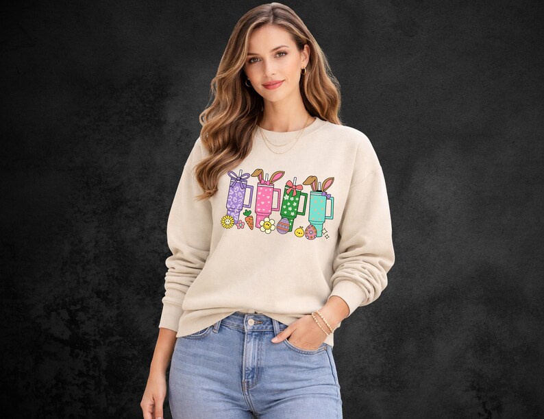 May include: Beige sweatshirt featuring a colorful graphic of Easter-themed mugs with bunny ears, carrots, and flowers. The design includes purple, pink, green, and blue mugs. The sweatshirt is worn by a person in blue jeans.