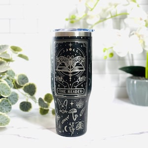 May include: Black stainless steel travel mug with a silver design. The design includes a moth, open book, and the text "THE READER." The mug has a silver rim and a blue lid. The design includes moons, stars, and other mystical symbols.