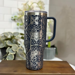 Puede incluir: Un vaso t&eacute;rmico azul marino con un dise&ntilde;o de mandala plateado y las palabras "F*ck Off" repetidas. Tiene un asa azul marino y una tapa transparente. El vaso est&aacute; sobre un soporte de madera.