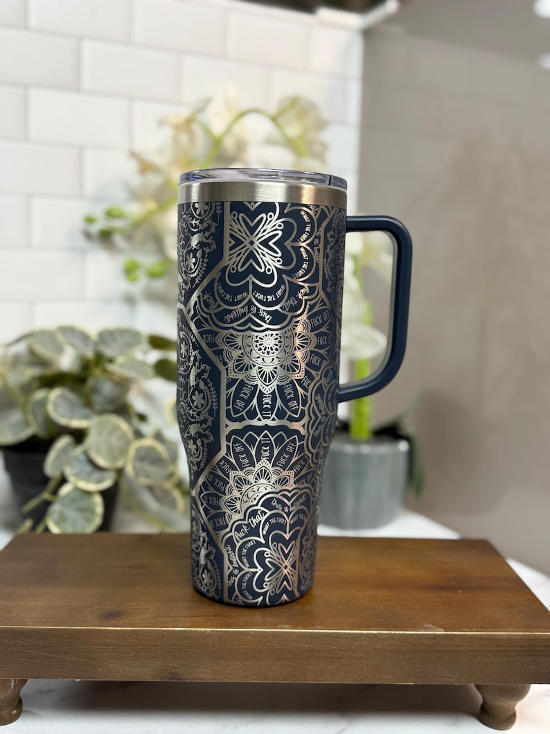 Puede incluir: Un vaso t&eacute;rmico azul marino con un dise&ntilde;o de mandala plateado y las palabras "F*ck Off" repetidas. Tiene un asa azul marino y una tapa transparente. El vaso est&aacute; sobre un soporte de madera.