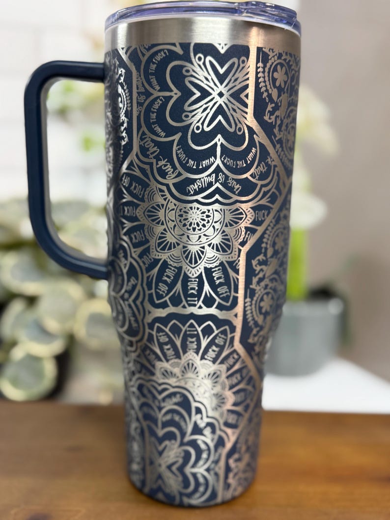 Puede incluir: Un vaso t&eacute;rmico azul marino con un dise&ntilde;o de mandala plateado y las frases "Fuck Off", "Fuck It" y "What the Fuck?" impresas. El vaso tiene un asa azul oscuro y una tapa transparente.