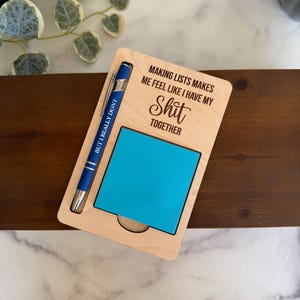 Può includere: Un portablocco in legno con un blocco note adesivo blu e una penna blu. Il portablocco ha il testo "MAKING LISTS MAKES ME FEEL LIKE I HAVE MY Shit TOGETHER" e la penna dice "BUT I REALLY DONT".