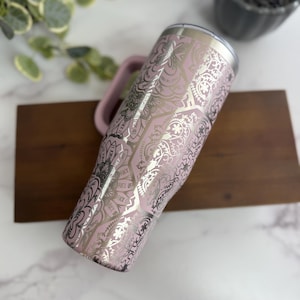 Puede incluir: Un vaso de acero inoxidable rosa con asa, con un dise&ntilde;o de mandala plateado y las palabras "FUCK OFF" en un patr&oacute;n repetido. El vaso tiene un borde plateado y un asa rosa.