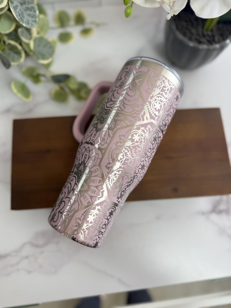 Puede incluir: Un vaso de acero inoxidable rosa con asa, con un dise&ntilde;o de mandala plateado y las palabras "FUCK OFF" en un patr&oacute;n repetido. El vaso tiene un borde plateado y un asa rosa.