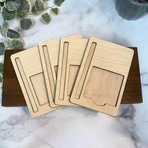 May include: Four light-coloured wooden phone stands with a rectangular design and a slot for the phone. The stands are arranged on a dark brown wooden base, with a marble background and greenery.