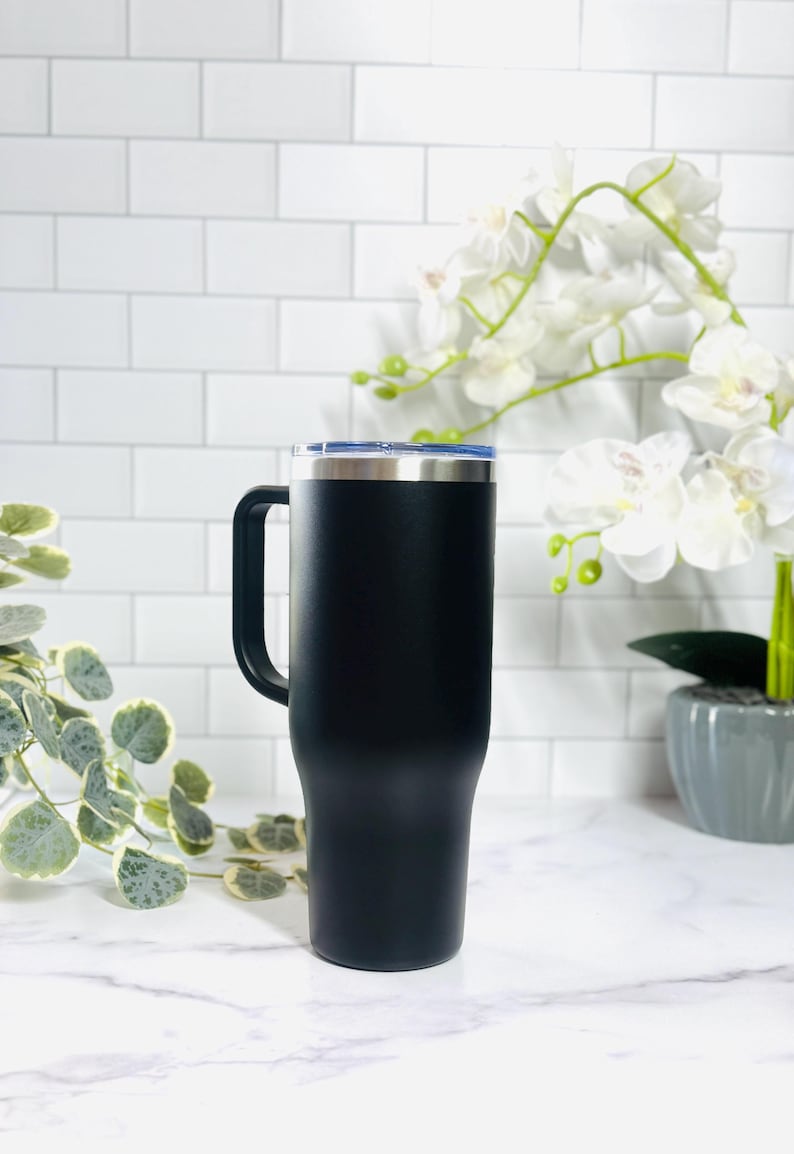 Puede incluir: Un termo de viaje negro con asa y borde plateado. El termo tiene una tapa azul y est&aacute; dise&ntilde;ado para mantener las bebidas calientes o fr&iacute;as. El termo es alto y cil&iacute;ndrico, adecuado para diversas bebidas.