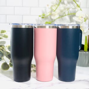 Puede incluir: Tres vasos aislados en azul marino, rosa y azul marino oscuro. Cada vaso tiene un borde plateado y una tapa transparente. Los vasos son cil&iacute;ndricos con una base ligeramente c&oacute;nica. El fondo es una pared de ladrillos blancos.