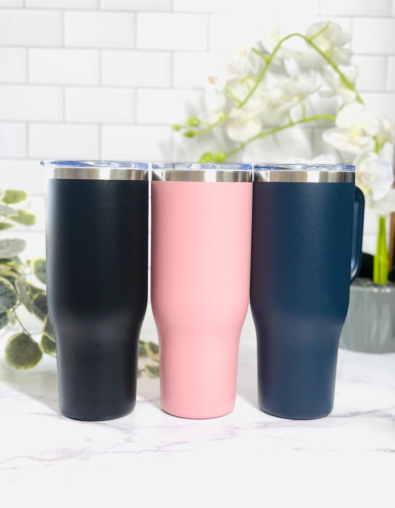 Puede incluir: Tres vasos aislados en azul marino, rosa y azul marino oscuro. Cada vaso tiene un borde plateado y una tapa transparente. Los vasos son cil&iacute;ndricos con una base ligeramente c&oacute;nica. El fondo es una pared de ladrillos blancos.