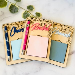 Może przedstawiać: Trzy spersonalizowane drewniane organizery na biurko z kwiatowymi akcentami. Każdy organizer zawiera notatnik i długopis. Imiona "Charlie", "Rachel" i "Hannah" są wyświetlane w kolorze niebieskim i różowym. Długopisy są niebieskie i różowe.