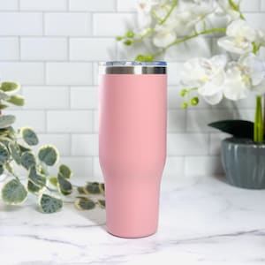 Puede incluir: Un vaso t&eacute;rmico rosa con borde plateado y tapa transparente. El vaso tiene un acabado mate y un dise&ntilde;o c&oacute;nico. Est&aacute; sobre una encimera de m&aacute;rmol blanco.