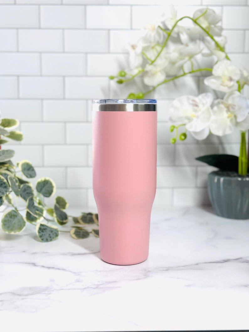 Puede incluir: Un vaso t&eacute;rmico rosa con borde plateado y tapa transparente. El vaso tiene un acabado mate y un dise&ntilde;o c&oacute;nico. Est&aacute; sobre una encimera de m&aacute;rmol blanco.