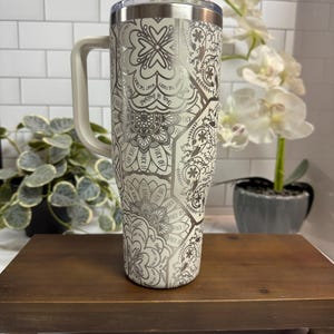 Puede incluir: Un vaso de acero inoxidable con asa, con un estampado floral y geom&eacute;trico color crema con las palabras "FUCK OFF" y "This is bullshit" impresas. El vaso tiene una tapa transparente.