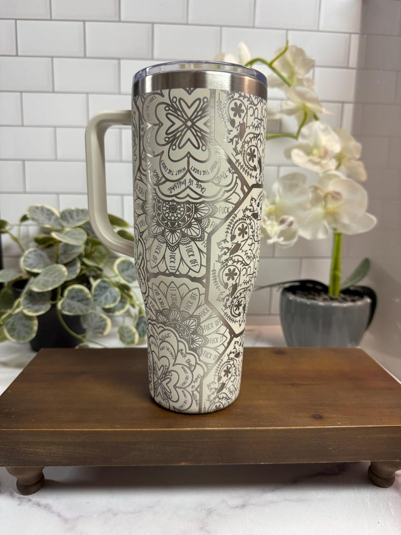 Puede incluir: Un vaso de acero inoxidable con asa, con un estampado floral y geom&eacute;trico color crema con las palabras "FUCK OFF" y "This is bullshit" impresas. El vaso tiene una tapa transparente.