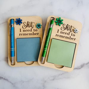 May include: Two wooden memo holders with pens. Each holder has a notepad, a pen, and the text "Shit I need to remember." One has a blue pen and notepad, the other a green pen and notepad. Decorative flower accents.