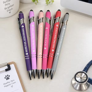 May include: A set of six retractable ballpoint pens in purple, light pink, peach, hot pink, red, and gray. Each pen features a silver clip, a black stylus tip, and white text with paw prints, including phrases like MORE FUR, LESS DRAMA.