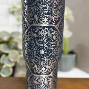 Puede incluir: Un vaso t&eacute;rmico azul marino con un patr&oacute;n floral y geom&eacute;trico plateado. El vaso tiene un borde y una tapa de acero inoxidable. El dise&ntilde;o incluye la palabra "Luck" en un patr&oacute;n repetido.