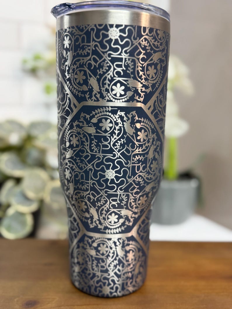 Puede incluir: Un vaso t&eacute;rmico azul marino con un patr&oacute;n floral y geom&eacute;trico plateado. El vaso tiene un borde y una tapa de acero inoxidable. El dise&ntilde;o incluye la palabra "Luck" en un patr&oacute;n repetido.