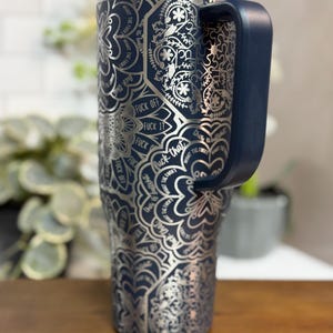 Puede incluir: Un vaso t&eacute;rmico azul marino con un dise&ntilde;o de mandala plateado y las palabras "FUCK OFF", "FUCK IT" y "FUCK THAT". El vaso tiene un asa azul marino y una tapa transparente. El vaso est&aacute; sobre una superficie de madera.