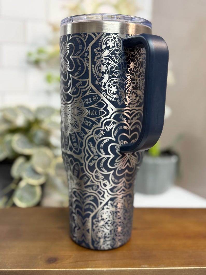Puede incluir: Un vaso t&eacute;rmico azul marino con un dise&ntilde;o de mandala plateado y las palabras "FUCK OFF", "FUCK IT" y "FUCK THAT". El vaso tiene un asa azul marino y una tapa transparente. El vaso est&aacute; sobre una superficie de madera.