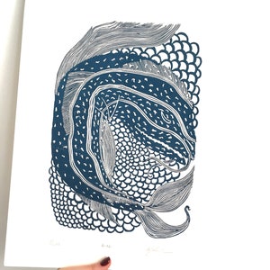 May include: A blue and white linocut print of a fish. The artwork features intricate patterns and textures, with a detailed depiction of the fish's scales and fins. The print is held by a hand against a white background.