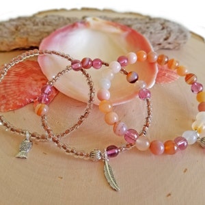 May include: Three beaded bracelets with pink, orange, and white beads. The bracelets have silver charms, one shaped like a fish and the other like a feather.
