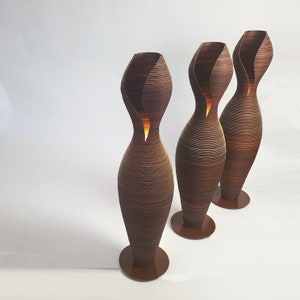 May include: Three brown, wooden, floor lamps with a unique, wavy, organic design. The lamps are lit and have a warm glow.
