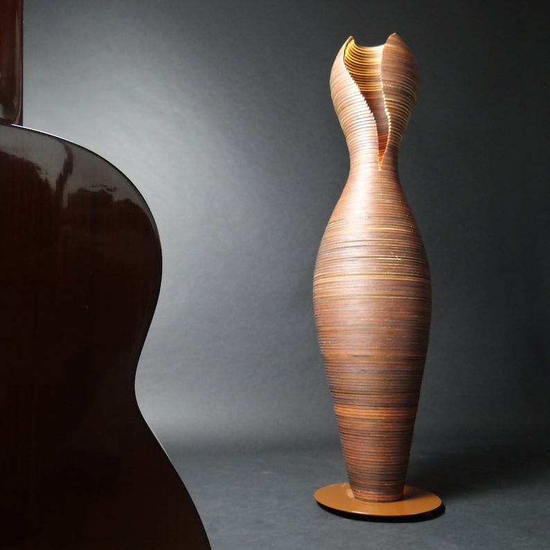 May include: A tall, brown, wooden vase with a unique, layered design. The vase has a narrow opening at the top and a wide base. The vase is standing on a brown base.