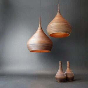 May include: Three wooden pendant lights in the shape of teardrops, with the two larger lights hanging from the ceiling and the smaller light sitting on a table. The lights are made of thin strips of wood that are layered and curved to create a teardrop shape. The lights are all lit and casting a warm glow.