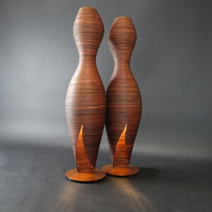 May include: Two tall, slender, wooden floor lamps with a warm, glowing light emanating from the bottom of each lamp. The lamps are made of thin, layered strips of wood, creating a unique and textured design.