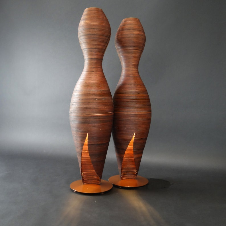 May include: Two tall, slender, wooden floor lamps with a warm, glowing light emanating from the bottom of each lamp. The lamps are made of thin, layered strips of wood, creating a unique and textured design.