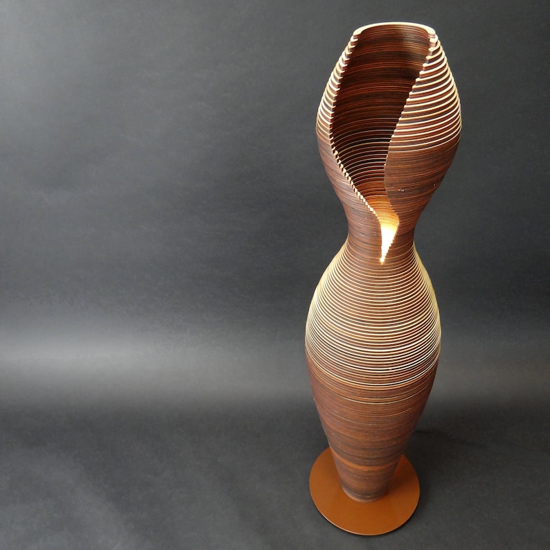 May include: A wooden floor lamp with a unique, layered design. The lamp is shaped like a vase with a wide opening at the top and a narrow base. The layers are made of thin strips of wood that are stacked on top of each other, creating a spiral effect. The lamp is lit from within, casting a warm glow.