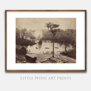 May include: Sepia-toned vintage print of a river scene with a steamboat, trees, and people on the shore. The print is framed in a brown wooden frame. The text "LITTLE WING ART PRINTS" and "VINTAGE AND DECORATIVE PRINTABLE ART" are at the bottom.