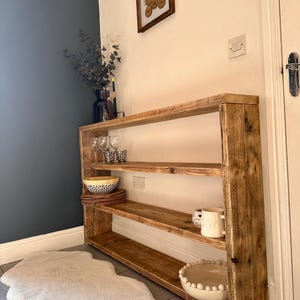 May include: A rustic wooden shelf unit with three shelves, featuring a natural wood finish. The shelf is filled with decorative items, including bowls, glasses, and a pitcher. The shelf is placed in a hallway with a white rug.