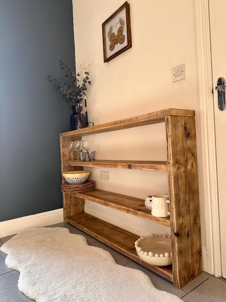 May include: A rustic wooden shelf unit with three shelves, featuring a natural wood finish. The shelf is filled with decorative items, including bowls, glasses, and a pitcher. The shelf is placed in a hallway with a white rug.