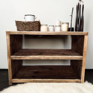May include: A rustic wooden shelf unit with two shelves. The shelf is made of reclaimed wood and has a natural finish. The shelf is decorated with candles, a basket, and a white pitcher.