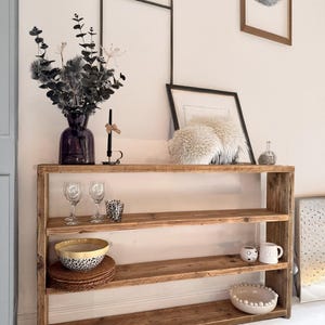 May include: A rustic wooden shelf unit with three shelves. The shelves are made of reclaimed wood and have a natural, weathered finish. The shelf unit is standing on a white floor and has a white wall behind it. There are various decorative items on the shelves, including a vase with dried flowers, glasses, bowls, and mugs.
