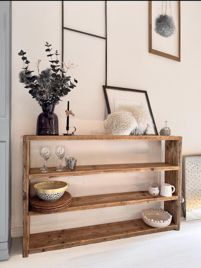 May include: A rustic wooden shelf unit with three shelves. The shelves are made of reclaimed wood and have a natural, weathered finish. The shelf unit is standing on a white floor and has a white wall behind it. There are various decorative items on the shelves, including a vase with dried flowers, glasses, bowls, and mugs.
