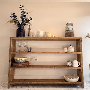 May include: A rustic wooden shelf unit with three shelves. The shelves are filled with various items, including a vase with dried flowers, glasses, bowls, and candles. The shelf unit is standing on a white tiled floor.