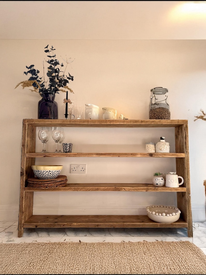 May include: A rustic wooden shelf unit with three shelves. The shelves are filled with various items, including a vase with dried flowers, glasses, bowls, and candles. The shelf unit is standing on a white tiled floor.