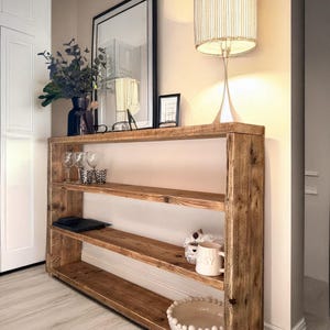 May include: A rustic wooden shelf unit with three shelves. The shelf is made of reclaimed wood and has a natural finish. The shelf is decorated with a lamp, a vase of flowers, and a bowl.