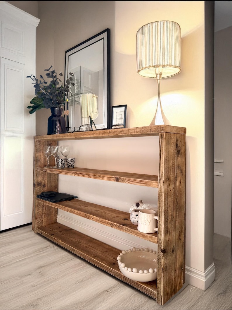 May include: A rustic wooden shelf unit with three shelves. The shelf is made of reclaimed wood and has a natural finish. The shelf is decorated with a lamp, a vase of flowers, and a bowl.