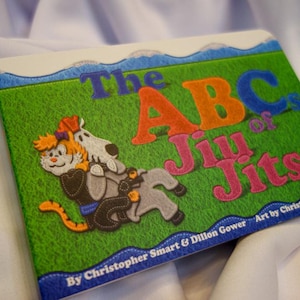 May include: A colorful book titled "The ABCs of Jiu Jitsu" with illustrations of a cat and a dog in a jiu jitsu pose. The book cover features a green background with blue, orange, pink, and white lettering. The text "By Christopher Smart & Dillon Gower" and "Art by Christopher Smart" are printed on the bottom of the cover.