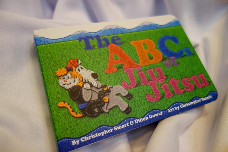May include: A colorful book titled "The ABCs of Jiu Jitsu" with illustrations of a cat and a dog in a jiu jitsu pose. The book cover features a green background with blue, orange, pink, and white lettering. The text "By Christopher Smart & Dillon Gower" and "Art by Christopher Smart" are printed on the bottom of the cover.