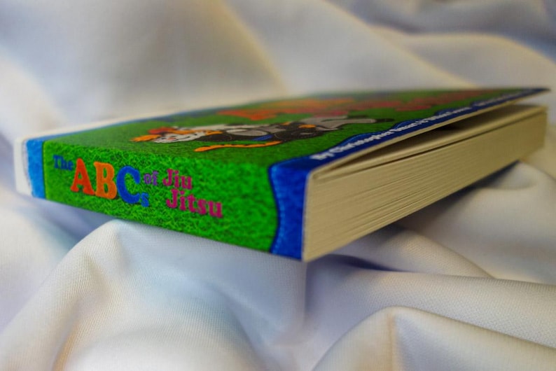 May include: A green, blue, and white book with the title "The ABCs of Jiu Jitsu" on the cover. The book is closed and lying on a white surface.