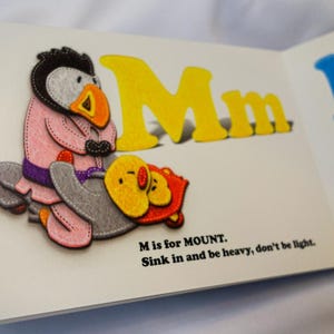 May include: A children's book page with a yellow felt letter M for Mount. The letter is accompanied by a felt illustration of a penguin wearing a pink jacket and a purple belt, holding a yellow chick. The text reads "M is for MOUNT. Sink in and be heavy, don't be light."