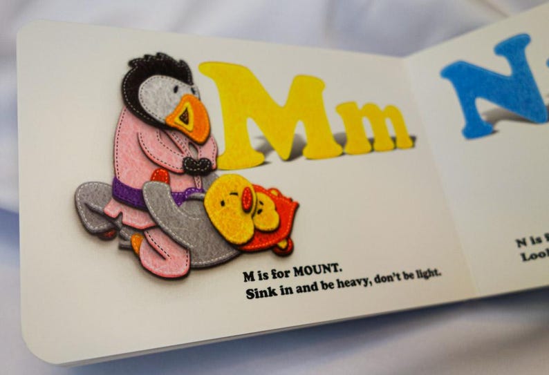 May include: A children's book page with a yellow felt letter M for Mount. The letter is accompanied by a felt illustration of a penguin wearing a pink jacket and a purple belt, holding a yellow chick. The text reads "M is for MOUNT. Sink in and be heavy, don't be light."