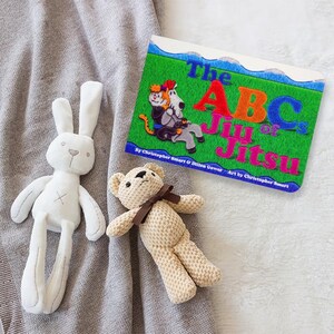 May include: A children's book titled "The ABCs of Jiu Jitsu" with colorful illustrations of animals practicing martial arts. The book is on a gray blanket with a white bunny and a brown teddy bear.