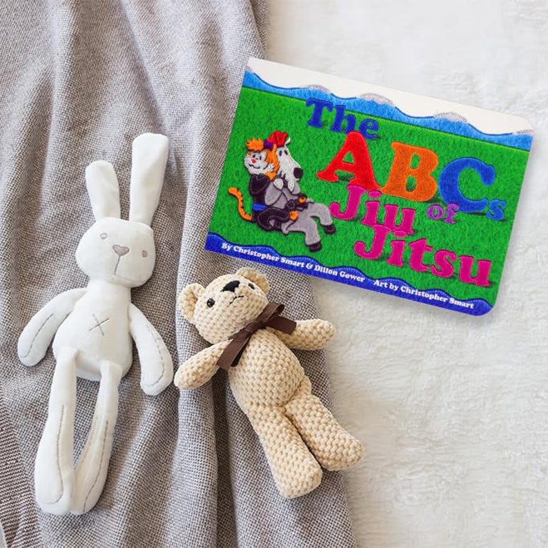 May include: A children's book titled "The ABCs of Jiu Jitsu" with colorful illustrations of animals practicing martial arts. The book is on a gray blanket with a white bunny and a brown teddy bear.