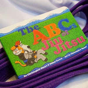 May include: A purple martial arts belt with a book titled "The ABCs of Jiu Jitsu" by Christopher Smart & Dillon Gower. The book cover features a cartoon illustration of a cat and a dog.