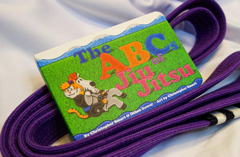 May include: A purple martial arts belt with a book titled "The ABCs of Jiu Jitsu" by Christopher Smart & Dillon Gower. The book cover features a cartoon illustration of a cat and a dog.